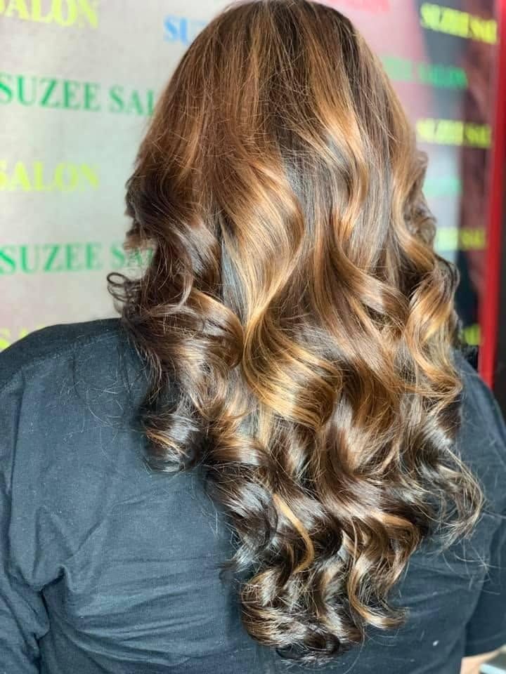 Rambut panjang keriting beralun, gelap dengan highlight tembaga keemasan, dilihat dari belakang di hadapan latar bercetak Suzee Salon